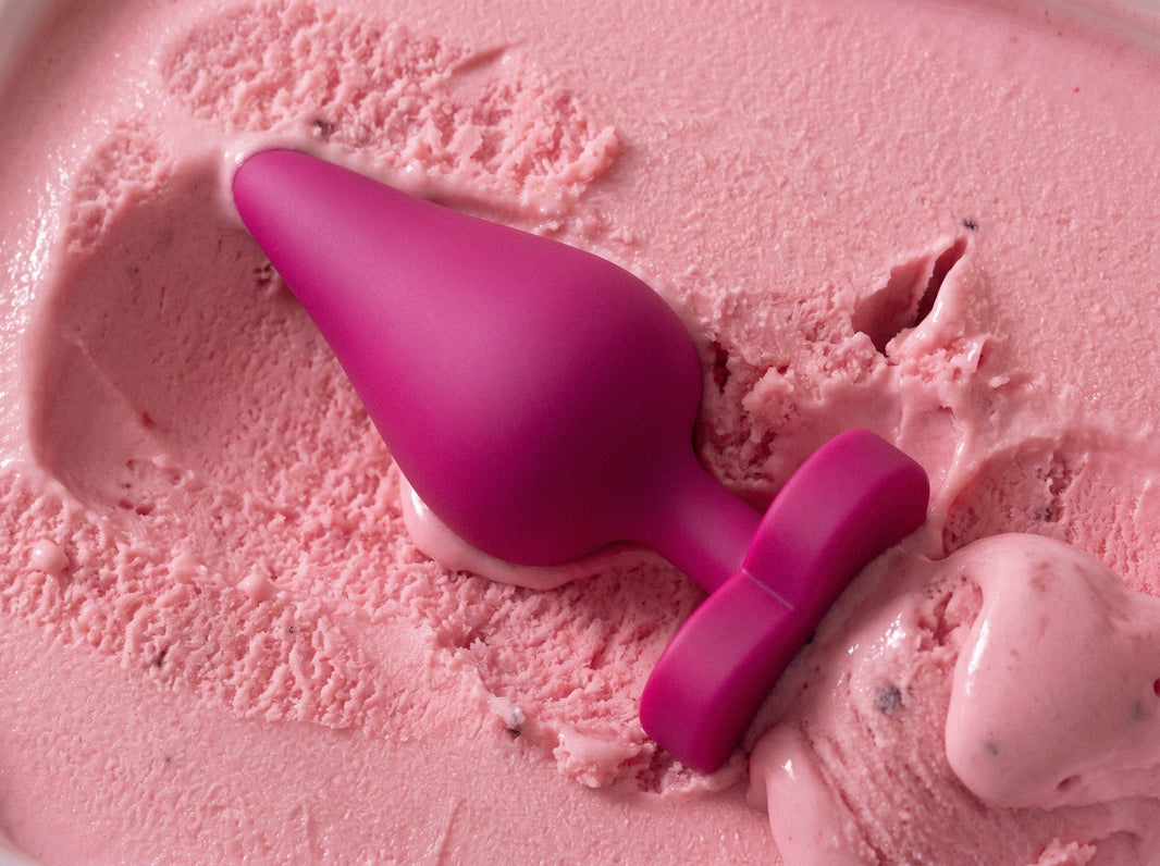 Magenta silicone butt plug with a tapered tip and wide base lying in a tub of pink ice cream