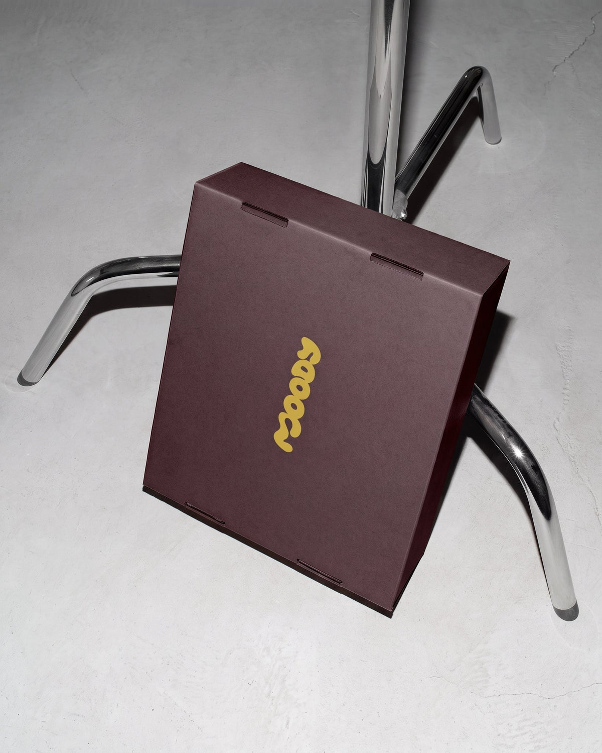 A dark brown rectangular product box with a gold vertical logo reading 'soops', photographed on a light gray concrete-like surface with a chrome stand behind it.