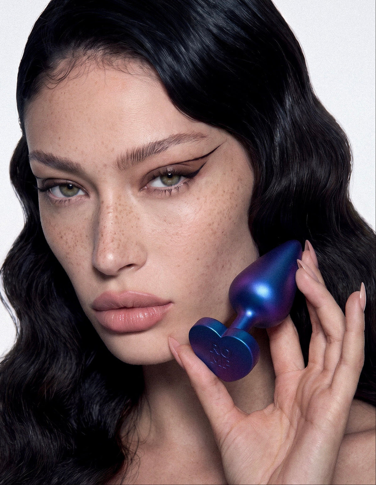 A close-up of a model with long dark wavy hair holding a metallic iridescent blue-purple intimate massager against her neck, set against a clean white background.