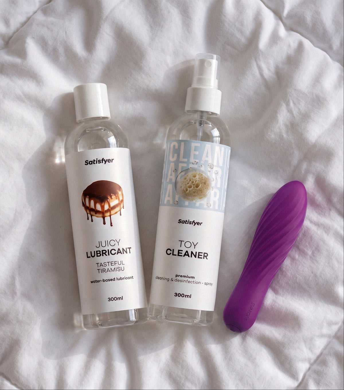 Two Satisfaction brand personal care products and a purple silicone toy are arranged on white quilted bedding. The products are a clear bottle of Juicy Lubricant with a white cap and label, and a clear spray bottle of Toy Cleaner with a white cap and label.