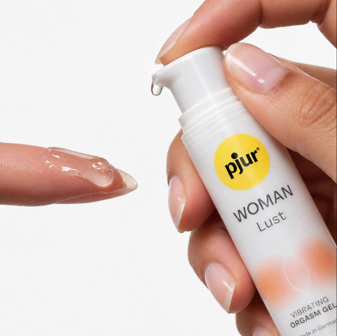 A close-up of a hand holding a white pump bottle labeled 'pjur WOMAN Lust' while applying clear gel to the tip of another finger against a plain white background.
