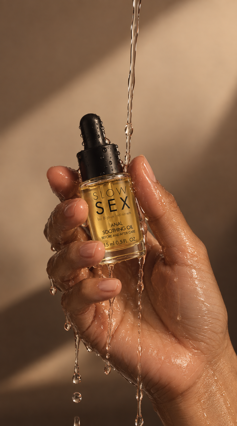 A hand holds a small glass dropper bottle labeled 'SLOW SEX' under running water, with warm sunlight casting shadows on a beige background.