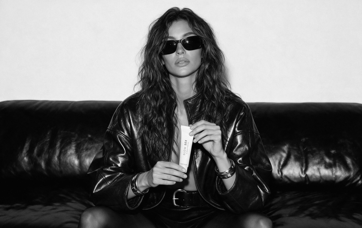 A black and white editorial photo of a model sitting on a black leather couch, wearing a shiny black leather jacket, sunglasses, and holding a white cosmetic tube.